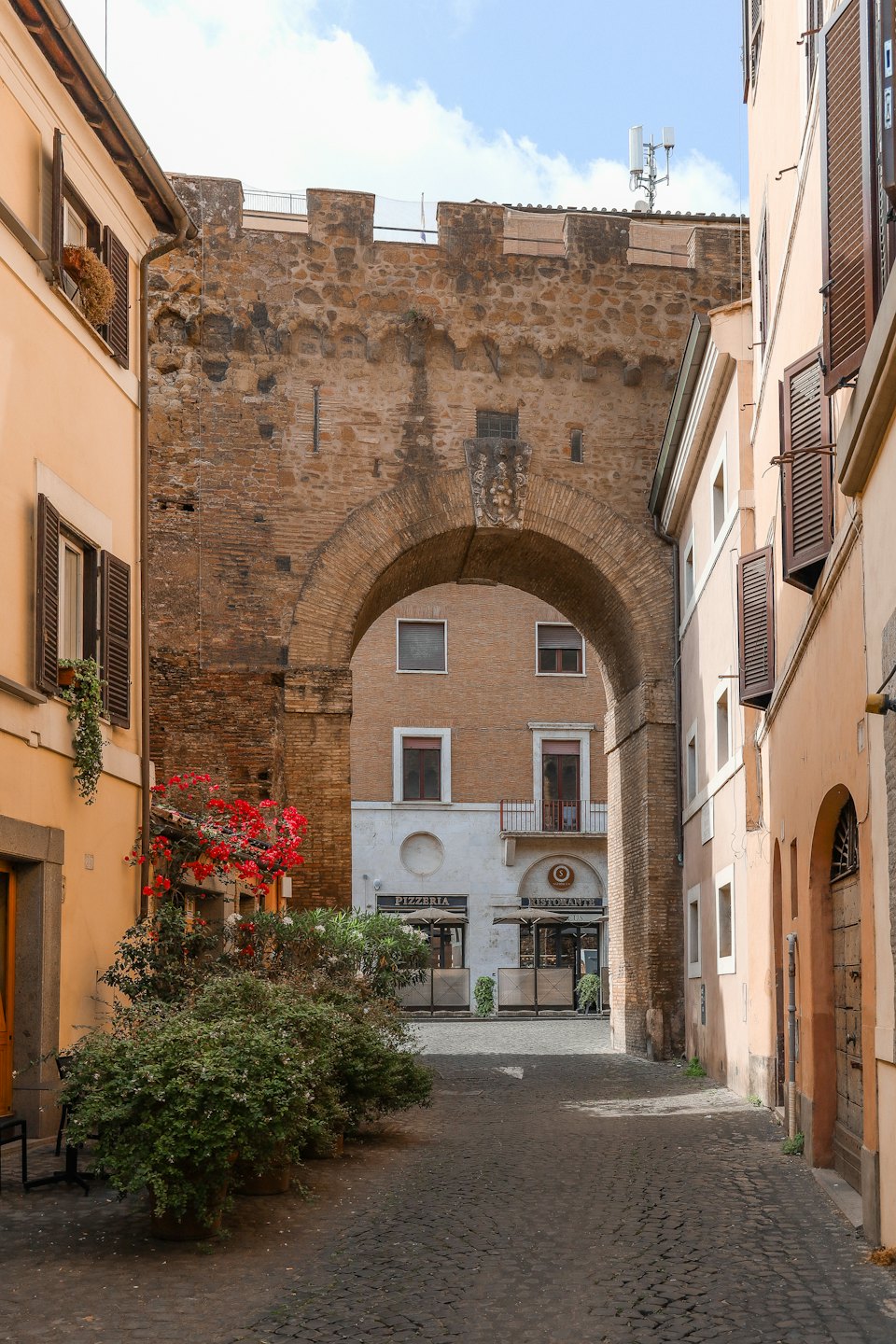 Strada acciottolata illuminata dal sole con edifici storici e insegna di pizzeria.